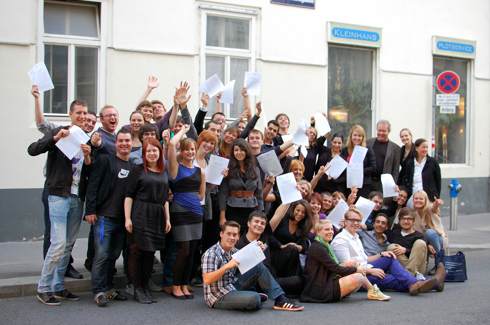 eine Gruppe von ca 50 Jugendlichen posieren stehend und sitzend für ein Foto und winken mit Papieren im A4 Format